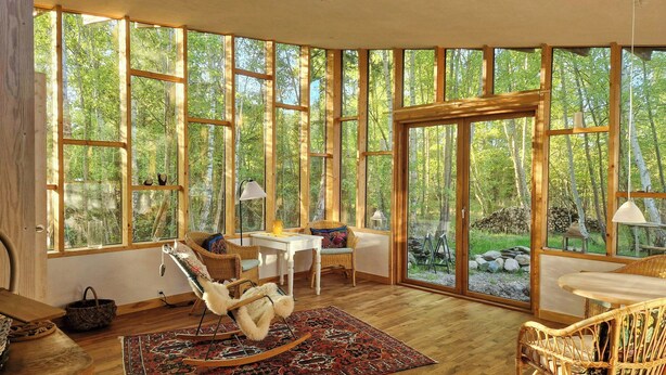 A room with large windows, a rocking chair, a table, and a wicker chair. - Hand-built timber and clay cottage by Bøtøskoven (Gedser)