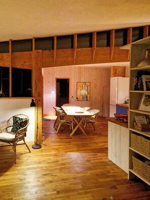 A dining area with a wooden table and chairs, a bookshelf, and a window with blinds. - Hand-built timber and clay cottage by Bøtøskoven (Gedser)