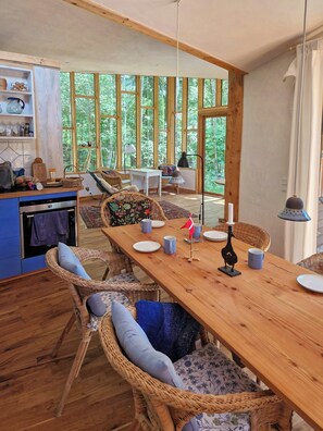 Una mesa de madera con sillas, una zona de cocina y ventanas grandes con vistas a un bosque.
