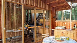 A wooden cabin with a dining area, a kitchen with a stove, and a large window overlooking greenery. - Hand-built timber and clay cottage by Bøtøskoven (Gedser)