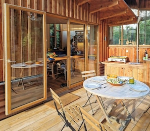 A wooden cabin with a dining area, a kitchen with a stove, and a large window overlooking greenery. - Hand-built timber and clay cottage by Bøtøskoven (Gedser)