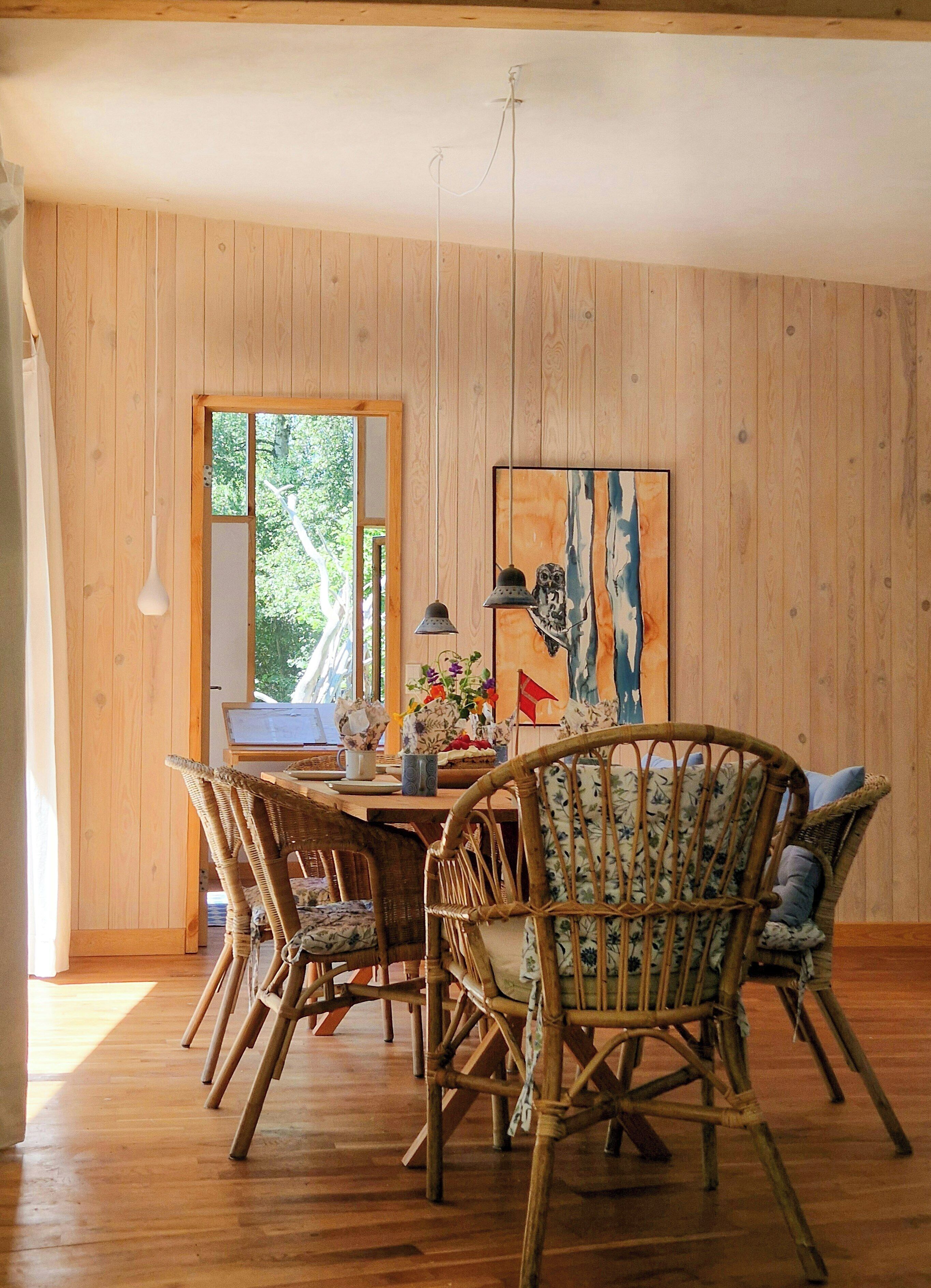 A dining area with a wooden table, wicker chairs, and a painting on the wall.