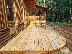 A wooden deck with a table and chairs, a wooden cabin with a large window, and a forested background.