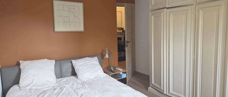 A bedroom with a bed, pillows, a bedside table with a lamp, a framed picture on the wall, and a wardrobe.