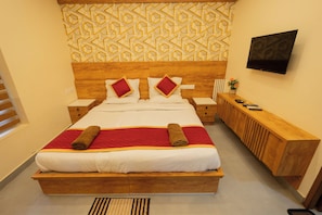 A hotel room with a large bed, wooden headboard, two bedside tables, a TV mounted on the wall, and a small wooden bench.