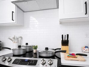 A modern kitchen with white cabinets, a stove, pots, a plant, and a cutting board.