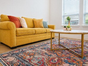 A yellow sofa with multiple cushions, a round table with a gold frame, and a patterned rug.