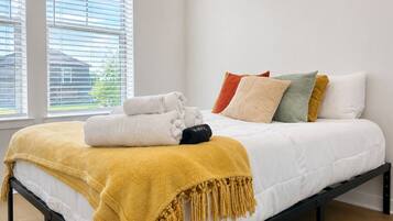 Una cama con colchón blanco, manta color mostaza y varias almohadas de distintos colores.
