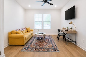 Uma sala de estar com sofá, mesa de centro, tapete, televisão e escrivaninha com cadeira.