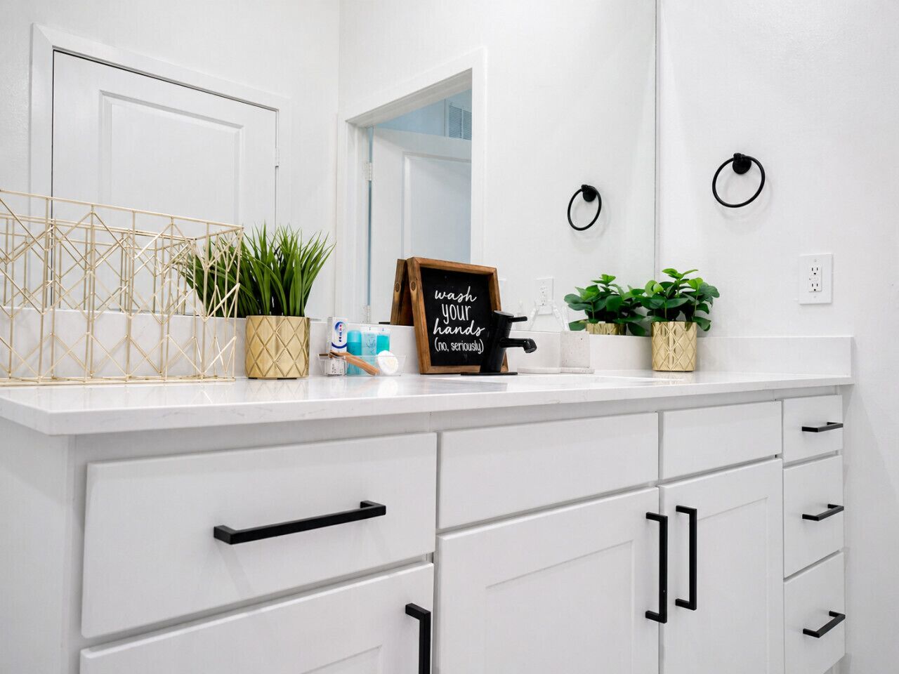 Un baño blanco con espejo, lavamanos, toallero y un cartel que dice 'lávate las manos (en serio)'.