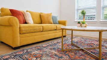 A yellow sofa with multiple cushions, a round table with a gold frame, and a patterned rug.