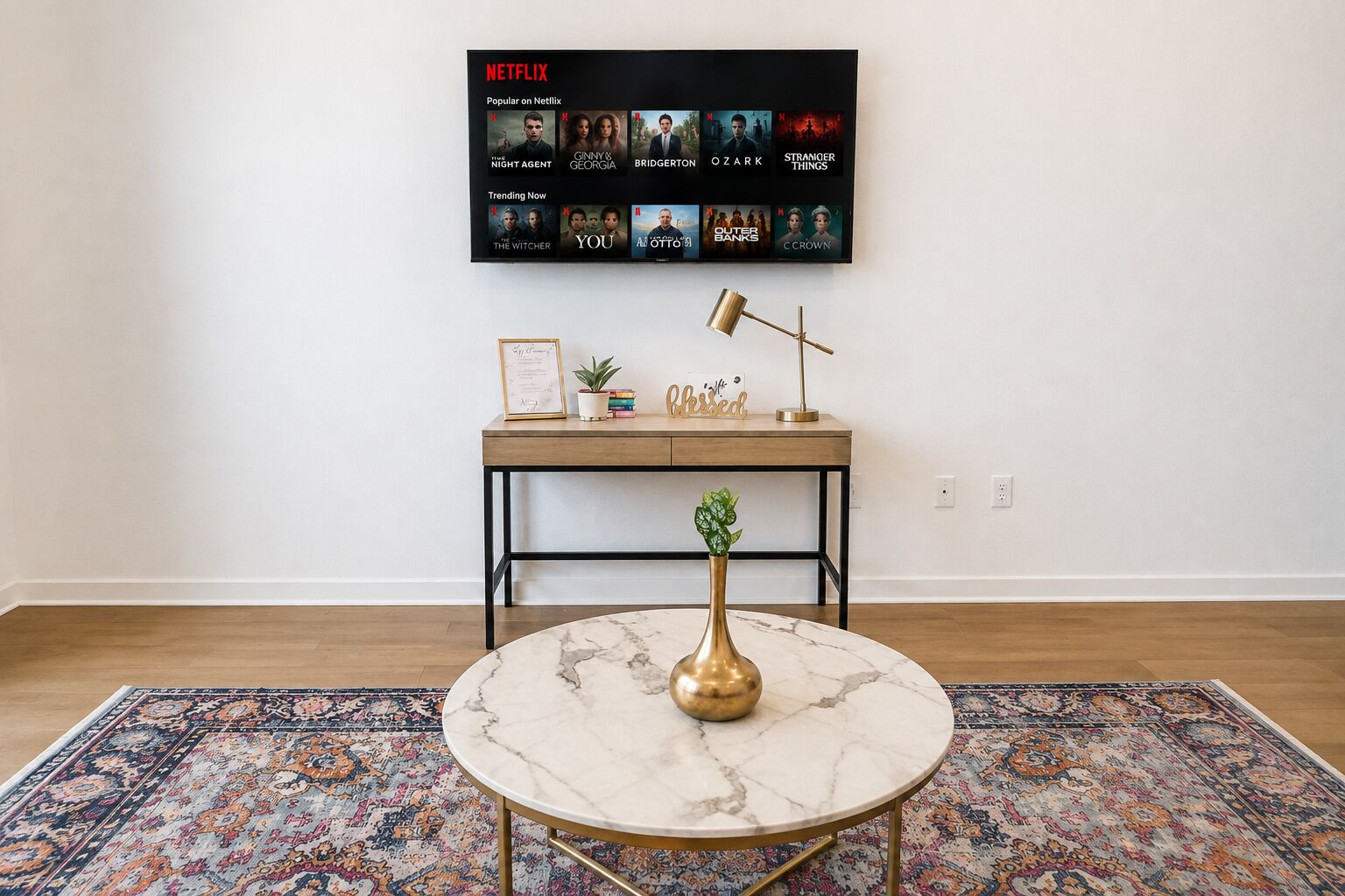 Una habitación con una mesa redonda de mármol, un escritorio de madera con un cuadro enmarcado, una planta en maceta y una pantalla de Netflix montada en la pared.