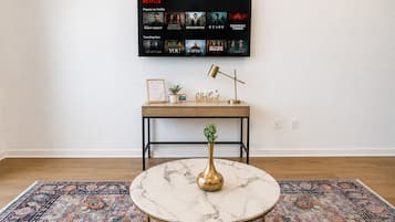 Una habitación con una mesa redonda de mármol, un escritorio de madera con un cuadro enmarcado, una planta en maceta y una pantalla de Netflix montada en la pared.
