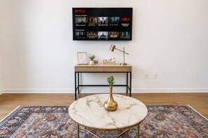 A room with a round marble table, a wooden desk with a framed picture, a potted plant, and a wall-mounted Netflix display.