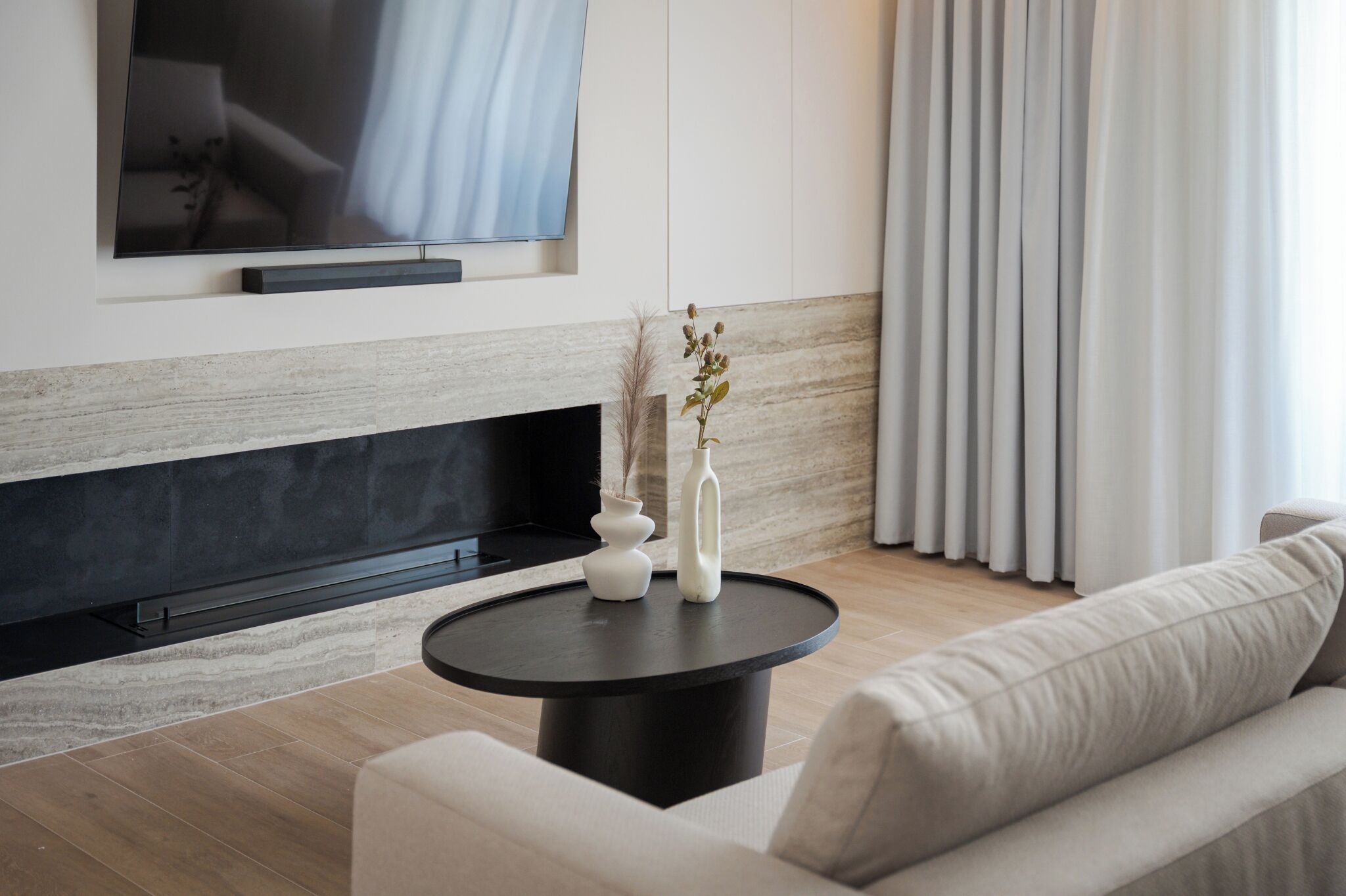 A modern living room with a flat-screen TV mounted on the wall, a black fireplace, a round coffee table with vases, and a sofa.