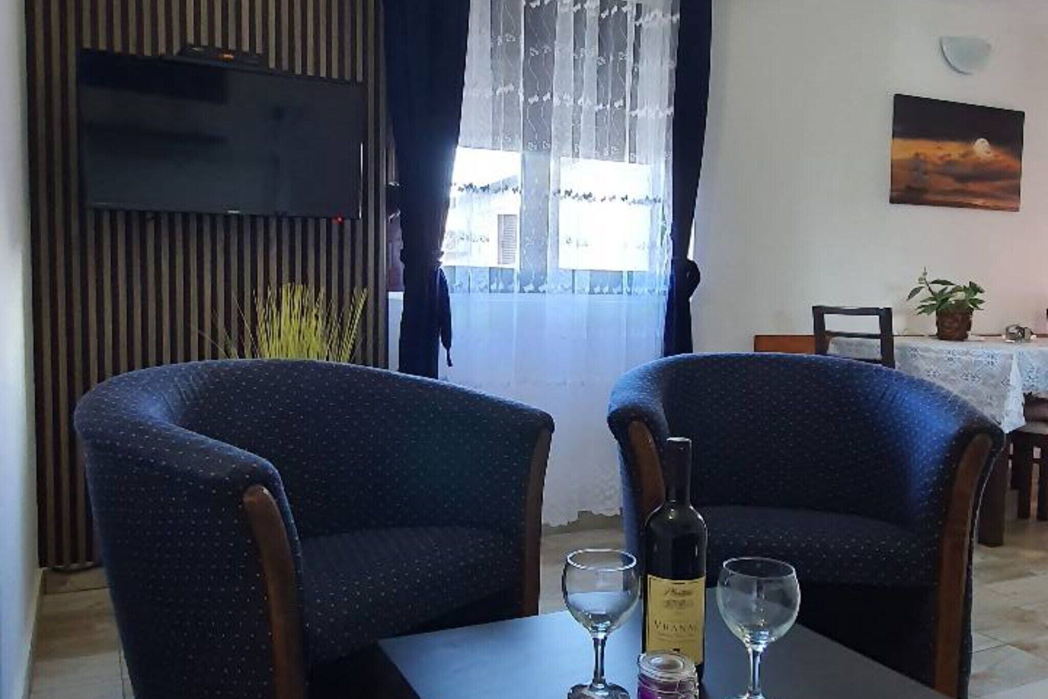Une pièce comprenant deux fauteuils, une table avec une bouteille et des verres, un téléviseur fixé au mur et une fenêtre avec des rideaux.