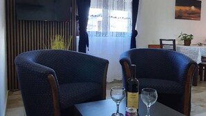 Huoneessa on kaksi nojatuolia, pöytä, jonka päällä on pullo ja laseja, seinälle asennettu televisio ja ikkuna, jossa on verhot.