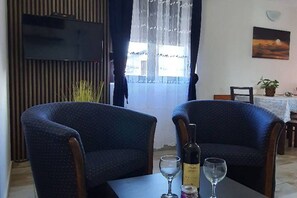 Ein Zimmer mit zwei Sesseln, einem Tisch mit Flasche und Gläsern, einem an der Wand befestigten Fernseher und einem Fenster mit Vorhängen.