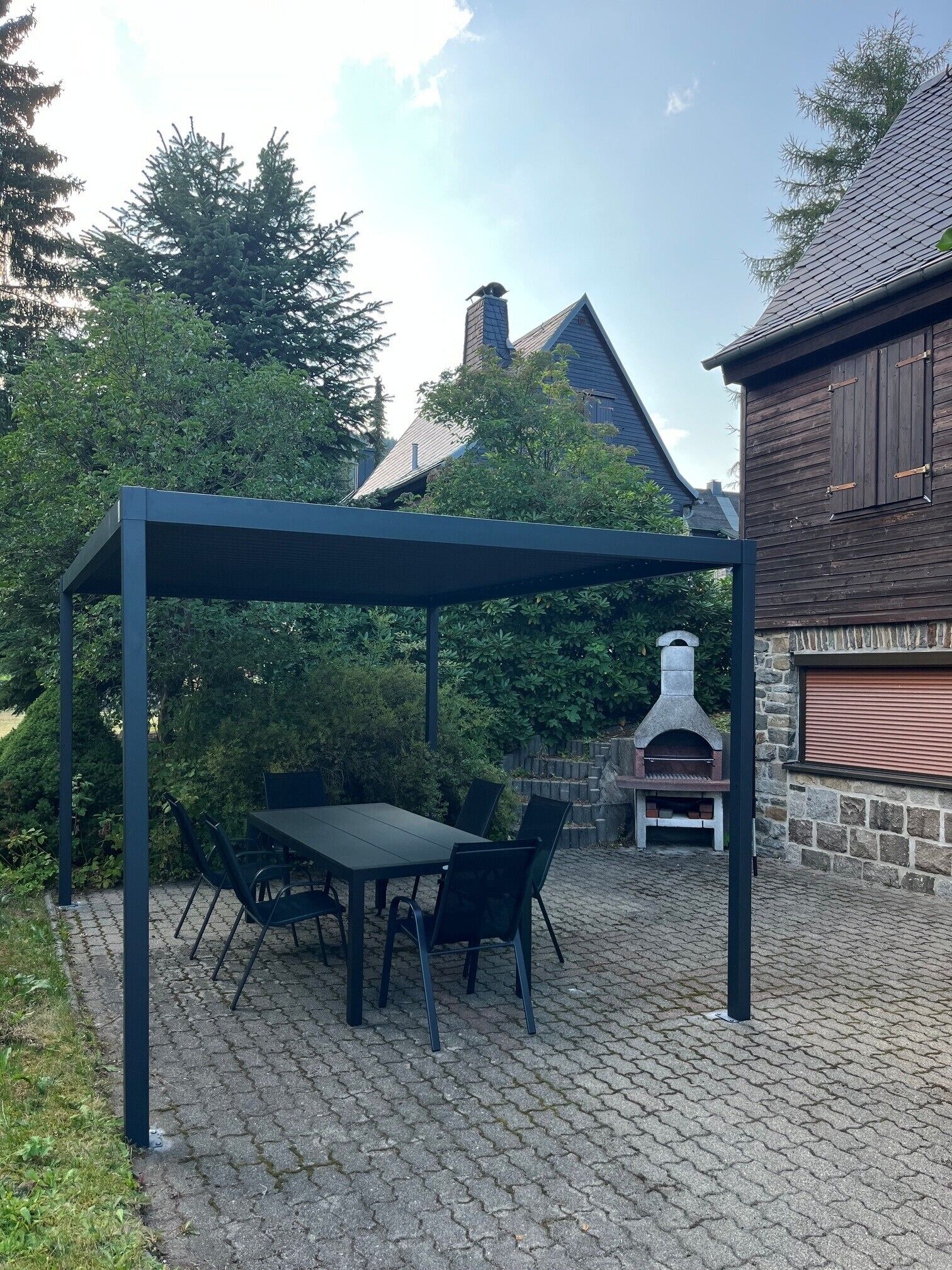 Ein überdachter Patio-Bereich mit Tisch und Stühlen, einem Steingrill und einer Steinmauer.