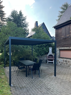 A covered patio area with a table and chairs, a stone barbecue grill, and a stone wall.