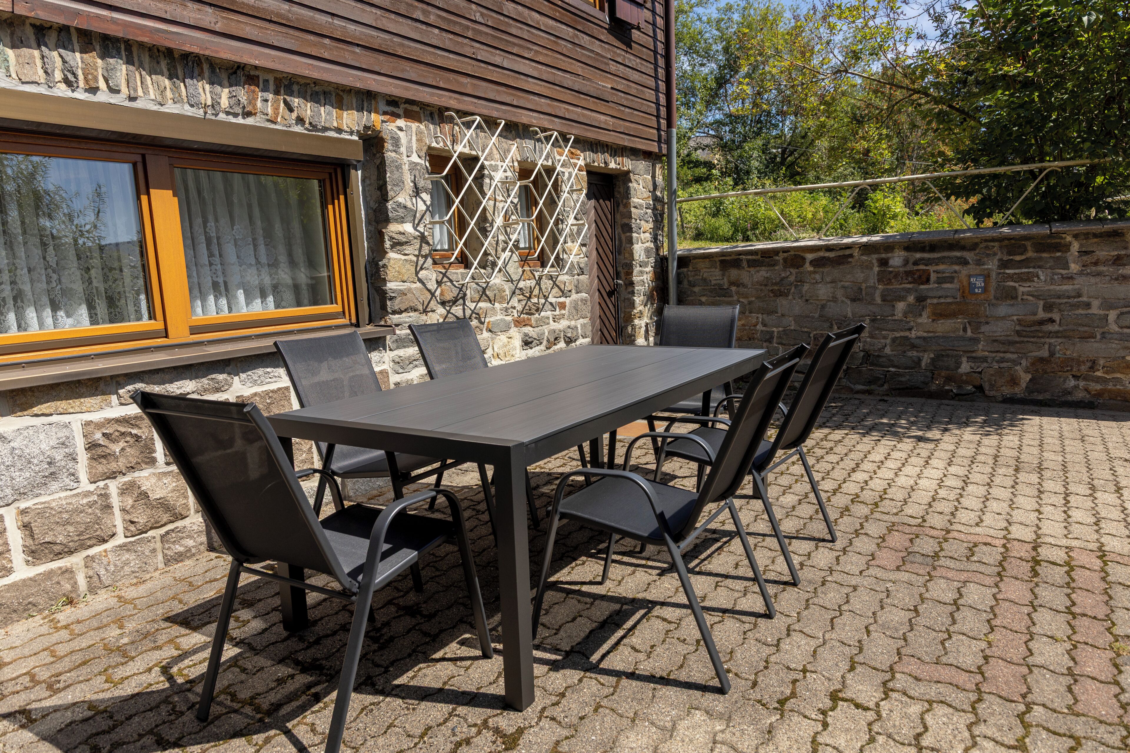 Eine Terrasse im Freien mit einem rechteckigen Tisch und sechs Stühlen, vor einer Steinwand mit Fenster und Tür.