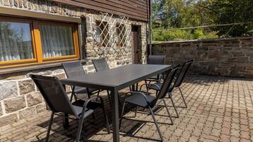 Eine Terrasse im Freien mit einem rechteckigen Tisch und sechs Stühlen, vor einer Steinwand mit Fenster und Tür.