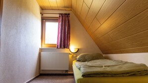 Habitació petita amb teulada inclinada, un llit individual, finestra amb cortines, radiador i làmpada penjada a la paret.