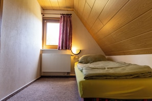 A small, sloped-roofed room with a single bed, a window with curtains, a radiator, and a wall-mounted light.