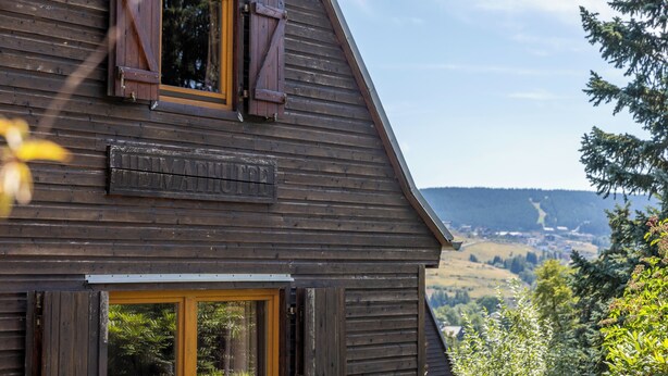 A wooden building with a window and a door, surrounded by greenery and a view of a valley.