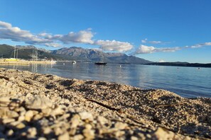 A pebble beach with calm water, boats, and mountains in the background. - Comfort Accommodation Baošići Montenegro-Double Room with Balcony and Sea View 2 (Baosici)