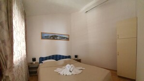 Hotellihuone, jossa on sänky, yöpöytä, lamppu, vaatekaappi ja taulu seinällä.