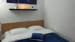 Valkoisilla lakanoiden ja sinisen tyynyn koristeltu sänky, jossa on puinen pääty ja seinälle kiinnitetty hylly, jossa on lamppu ja koriste-esineitä.