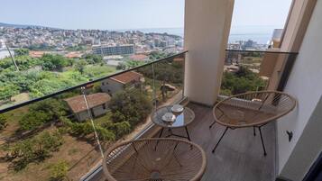 A balcony with two wicker chairs and a small table, overlooking a cityscape with buildings and greenery.