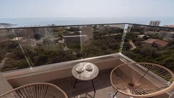 A balcony with a glass railing, two wicker chairs, a small round table, and a view of the sea and buildings.