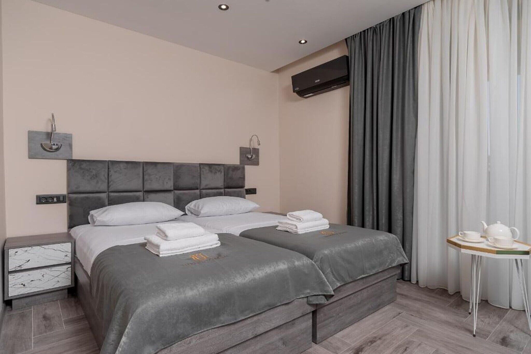 Ein modernes Hotelzimmer mit einem großen Bett, Nachttischen, einem kleinen Tisch mit Teekanne und einem Fenster mit Vorhängen.