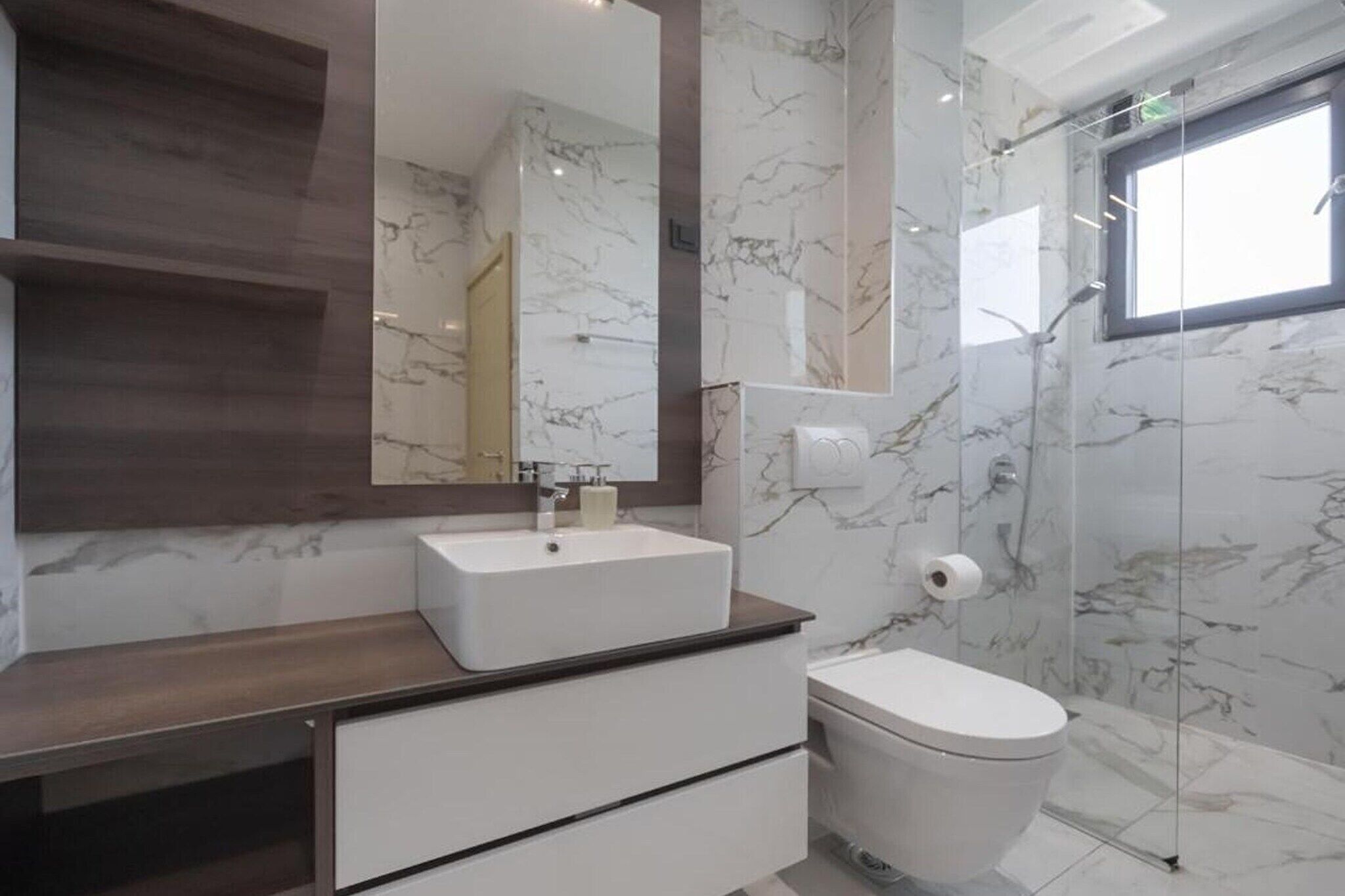 A modern bathroom with a marble wall, a large mirror, a white sink, and a toilet.