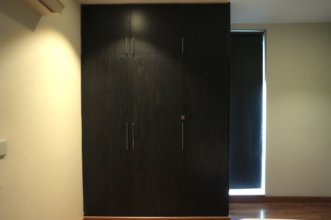 A dark wooden wardrobe with two handles and a small lock, positioned in a room with a plain wall and a window.