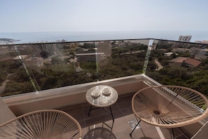 Un balcó amb barana de vidre, dues cadires de vimet, una taula rodona petita i vista al mar i edificis.