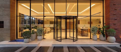 Entrada de hotel com porta giratória, plantas em vasos e uma área de recepção visível através das portas de vidro.