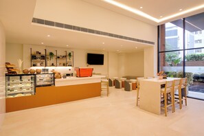 A modern cafe interior with a counter displaying pastries and a coffee machine, seating area with tables and chairs, and a large window offering a view of the cityscape. - Lemon Tree Premier Vadodara (Vadodara)