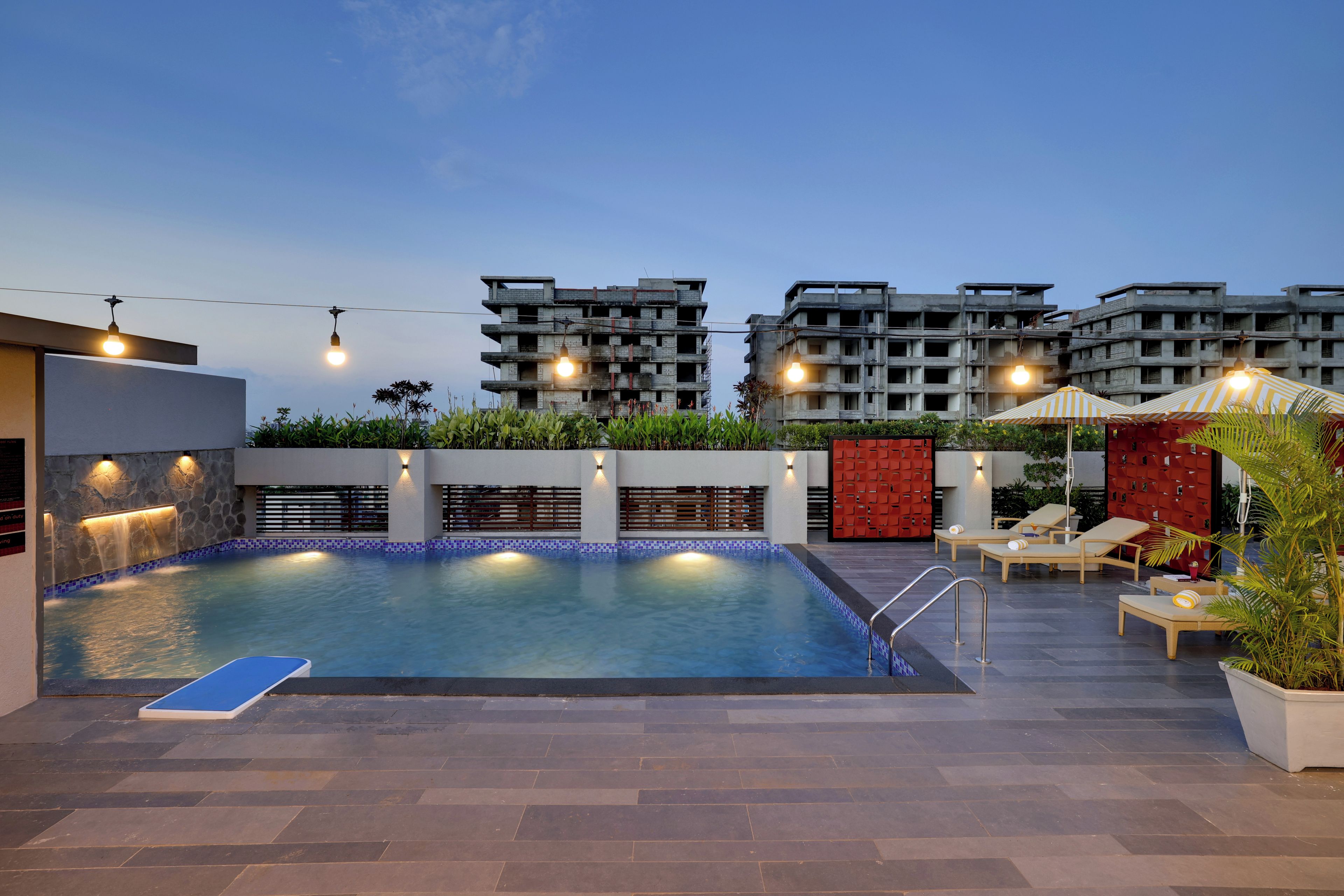 A rooftop swimming pool with a blue tiled edge, surrounded by lounge chairs and a red wall.