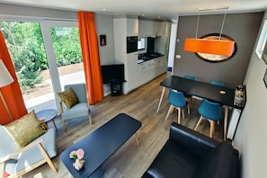 A modern living room with a sofa, a dining table with chairs, a kitchen area, and a large window with curtains.