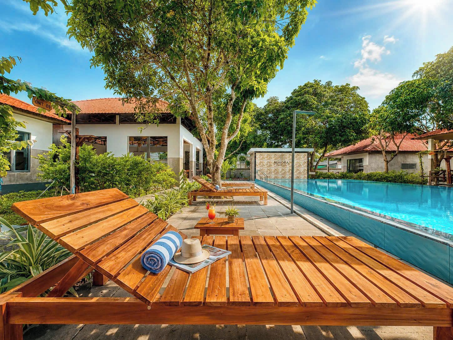 Un espace aménagé au bord de la piscine, avec des chaises longues en bois, une piscine et un bâtiment portant l’inscription « AURORA ».
