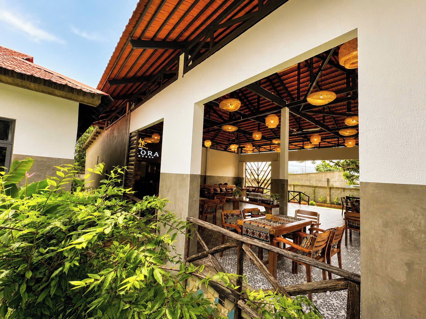 Un área de comedor al aire libre con mesas y sillas de madera, un espacio de estar cubierto con luces colgantes y la entrada de un edificio con un letrero.