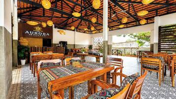An indoor dining area with wooden furniture and patterned tablecloths, a visible restaurant sign, and a tiled floor.