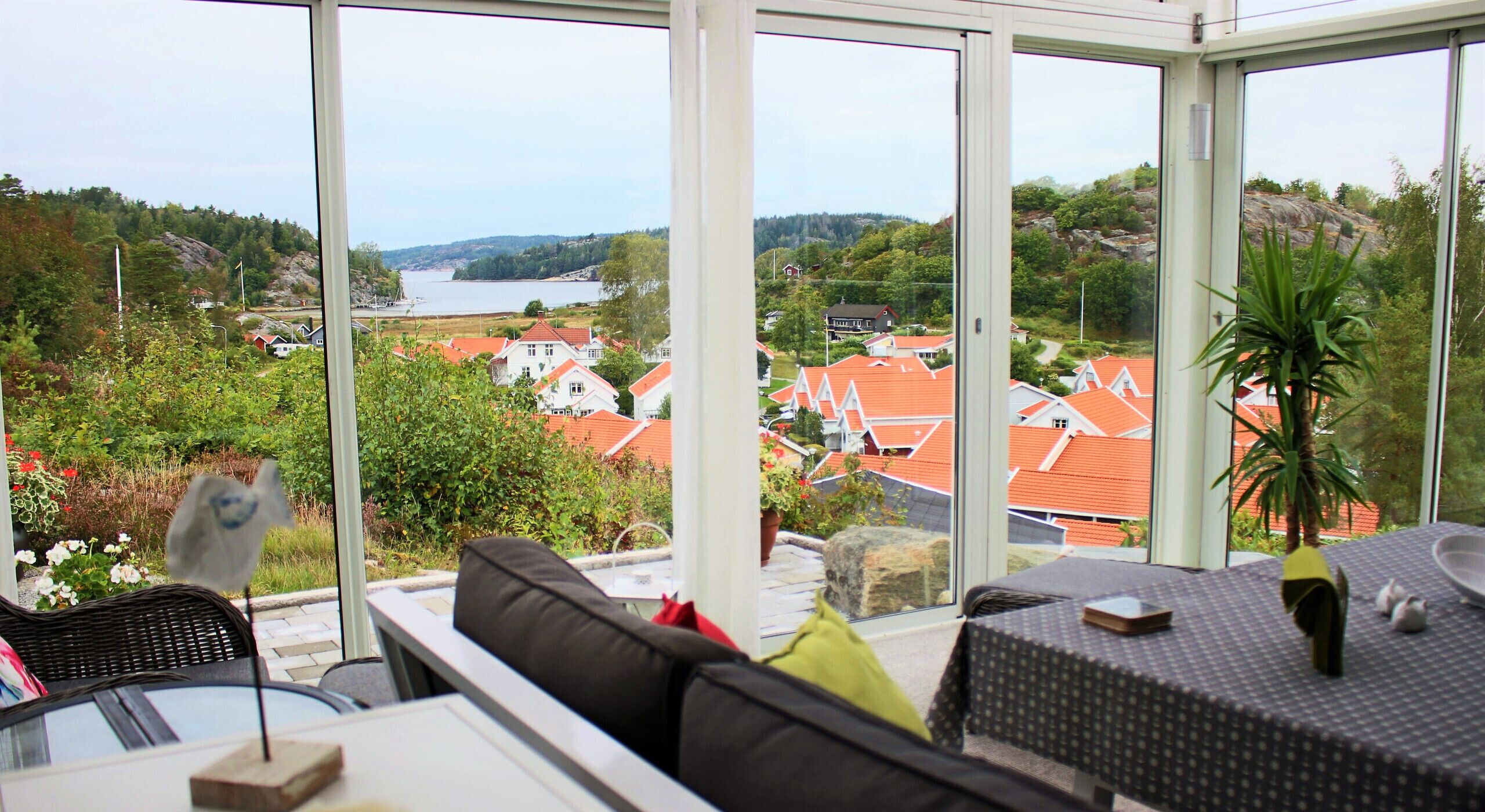 A conservatory with a view of a coastal village, furnished with a sofa and a dining table.