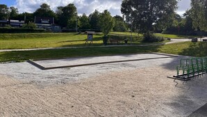 Uma área pavimentada com um banco, um campo gramado e árvores ao fundo.