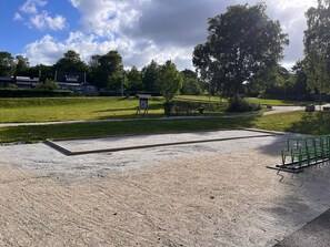 Ein gepflasterter Bereich mit einer Bank, einer Wiese und Bäumen im Hintergrund.