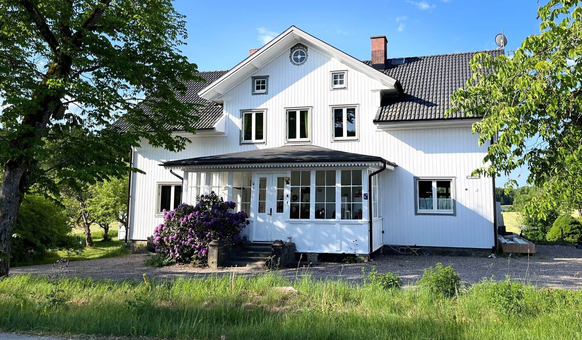 A white two-story house with a covered porch, surrounded by greenery and trees.