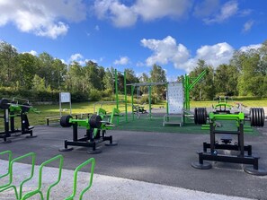 Ein Outdoor-Fitnessbereich mit unterschiedlichen Trainingsgeräten, einem Hinweisschild und einer Rasenfläche.